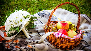 basket of fruits beside bouquet of white petaled flowers on top of gray floral blanket